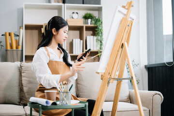 Asian Female painter do artwork in art workshop, painting supplies, oil pastels, two canvas easel, creative space with paintbrush in art studio