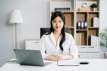 Young Asian woman smiling thinking planning writing in notebook, tablet and laptop looking at camera at office