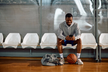 Happy black athlete texting on mobile phone on basketball court.
