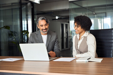 Two busy happy diverse professional business people working in office with laptop computer. Middle aged male executive manager talking to young female colleague having conversation sitting at work.