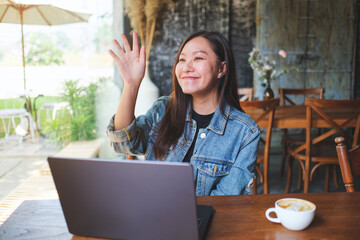 Portrait image of a woman raising hand and greeting to colleague while working on laptop computer in cafe