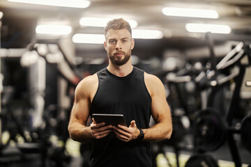 Portrait of a fitness instructor standing in a gym with tablet in his hands and looking at the...