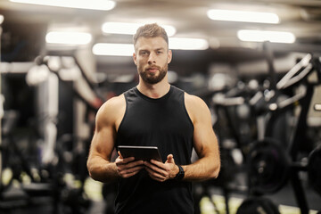 Portrait of a fitness instructor standing in a gym with tablet in his hands and looking at the camera.