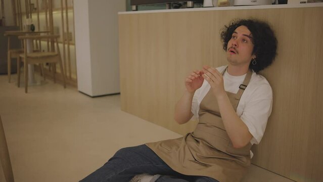 A guy waiter, tired from overwork, sit on the floor in a cafe next to the bar counter and upset. Frustrated man sits on the floor