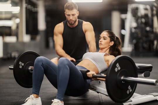 A sportswoman is doing barbell hip thrust in a gym and her personal trainer is helping her.