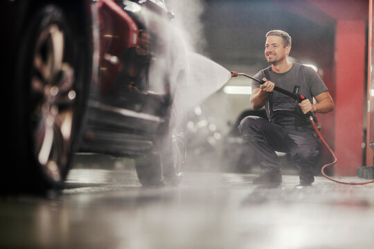 Smiling carwash serviceman washing car with high pressure water gun at station.