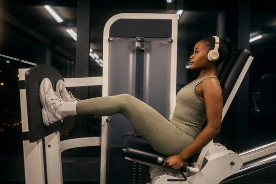 Side view of an african american sportswoman with headphones sitting on a leg press machine and doing workouts at gym.