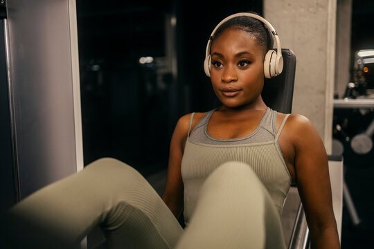 Front view of black sportswoman in shape with headphones doing leg press workouts at gym.