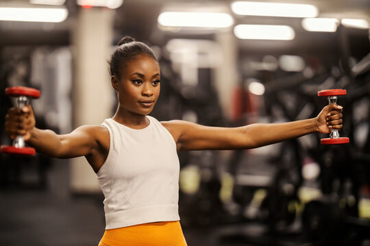 A fit black sportswoman doing workouts with dumbbells and practicing her shoulders at gym.