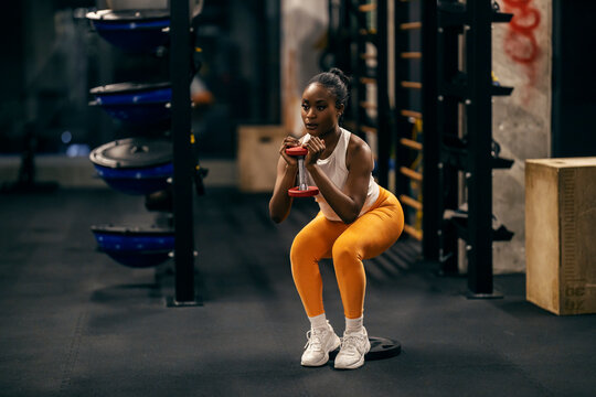 A strong black sportswoman in squatting positing practicing with dumbbells at gym.
