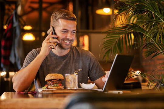 Smiling man having phone call and using tablet during his lunch break at restaurant.