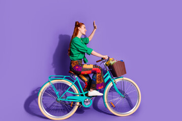 Full size photo of nice woman dressed green shirt pants riding bike waving hand look empty space isolated on purple color background