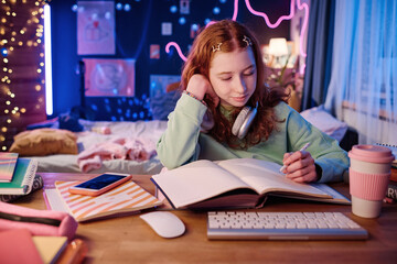 Medium shot of Caucasian girl with red hair sitting at desk in her room doing school homework in evening, copy space
