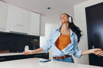 Happy woman enjoying music in modern kitchen with arms outstretched while standing on counter
