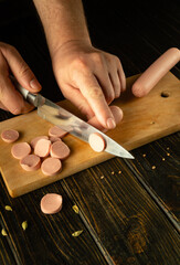 Slicing vienna sausage on a cutting board with a knife in a chef hand for preparing a dish for lunch. Space for advertising on a dark background