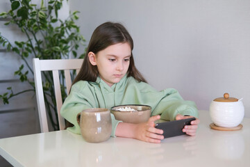 Girl of 8 or 9 years old uses mobile phone while having breakfast on table. Child dictates his own...