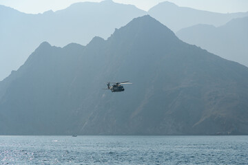 The helicopter soars gracefully over the calm waters while the mountains of Oman provide a...