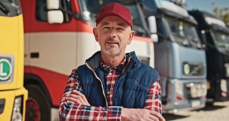 Portrait of mature truck driver standing outside on background of truck cars. Adult man looking at camera and posing with crossed arms. Concept of ordinary professions. Worker at workplace.