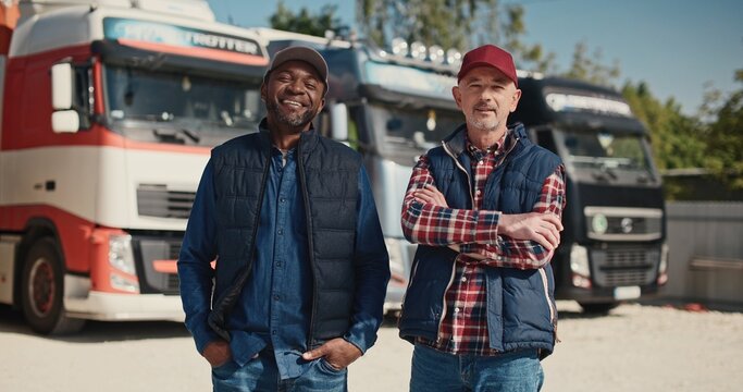 Two mixed-race coworkers standing before large trucks outside. Mature drivers in hats and jackets positively looking into camera. Getting ready to traveling. Working together in delivery company.