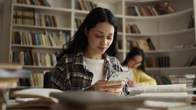 Smiling young woman hold smartphone scrolling social media online while take a break at university library Pretty student relax enjoying great day after lessons indoors