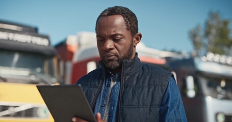 Professional truck driver. African American trucker checking his route on tablet computer with dispatcher and standing at background of long vehicle. Transportation service.