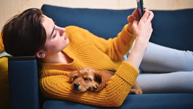 A woman is lying on the sofa with her pet and using a smartphone, browsing the Internet on her phone in the living room