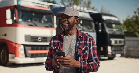 Good-looking African American man typing message on his smartphone while standing at trucks parking. Using his gadget for work. Remote communication. Working at distribution center.