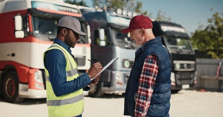 Side view of two multiethnic workers speaking with each other about load and unload freight at warehouse. Actively speaking while standing at lorries parking in sunny day. Shipping concept. - obrazy, fototapety, plakaty