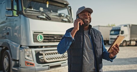 African American man speaking on phone with his coworker or customer while holding tablet device. Busy worker standing at trucks parking while remotely communicating. Concept of technology.