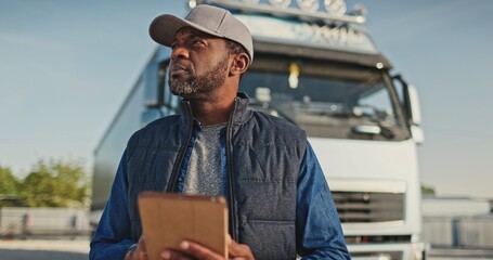 Handsome African American man using his tablet device while standing next large truck. Typing...