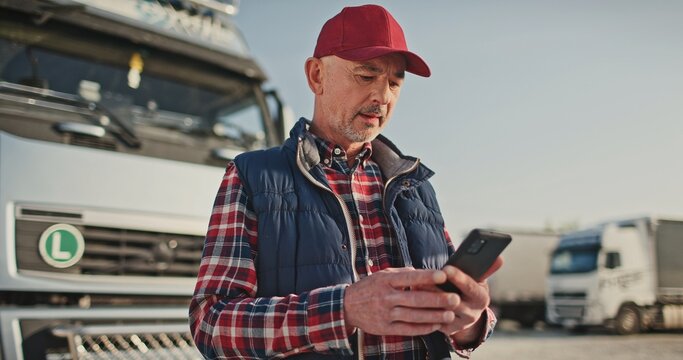Busy Caucasian male driver chatting on his smartphone with someone about agreement on place of future delivery while standing in front of large trucks with merchandise. Transport concept.