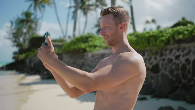 Smiling handsome guy with a mobile phone taking picture of azure waters of the Pacific ocean from fascinating beach, enjoying vacation on tropical island with exotic nature. High quality 4k footage