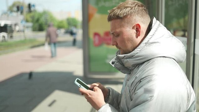 Young attractive bearded Caucasian man young adult male sit at public transport bus station waiting with mobile smart phone with backpack student or tourist city life 