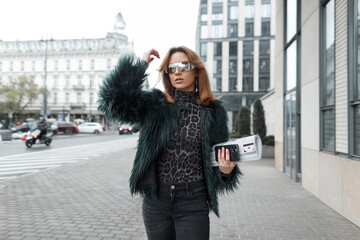 Glamorous beautiful fashionable woman with sunglasses in a fashion shaggy jacket with a magazine and phone walks in the city