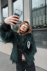 Stylish beautiful fashion woman model with sunglasses in a fashionable green shaggy jacket with a magazine takes a selfie photo on a smartphone in the city