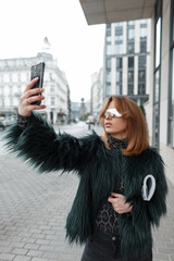 Fashion beautiful young girl with sunglasses in a stylish jacket takes a selfie on a smartphone in the city