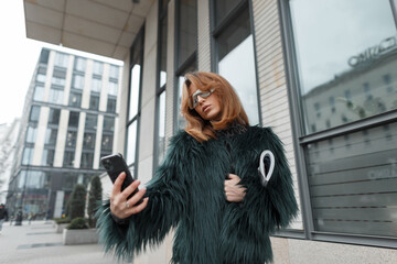 Beautiful fashionable young woman model in fashion clothes with a shaggy jacket with a magazine takes a selfie photo on a smartphone in the city