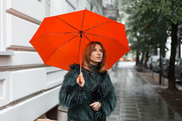 beautiful fashion young girl in fashionable clothes with a bright orange umbrella walks in the city on a cloudy rain day