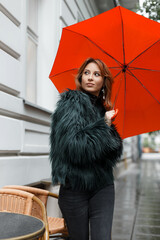 A bright beautiful fashion woman in stylish clothes with an orange umbrella walks in the city on a rainy day