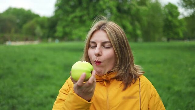 Close up portrait of attractive Caucasian woman eat green apple fruit outdoors at city spring park. Beautiful active female stand with happy face, enjoy eating apple, healthy foods for health care