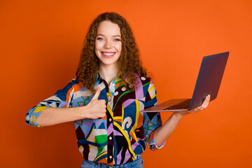 Photo of lovely cheerful girl teenager wear stylish print clothes wireless device thumb up gesture isolated on orange color background