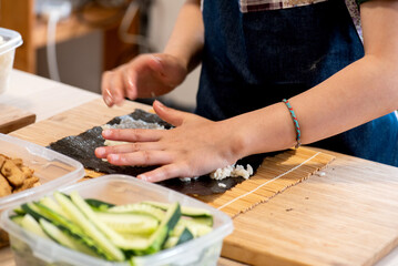 detail of girl's hands making sushi, squeezing rice on nori seaweed, in front of lunch box with sliced cucumber pieces ready for the little girl to cook on her own