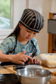 Girl making sushi rolls, dressed in denim apron and chef's hat. natural light, window behind with nature in the background.