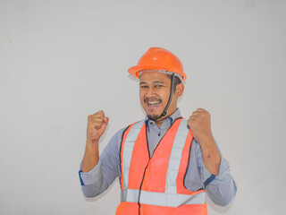Asian man worker wearing safety helmet lookis happy celebrating his victory by clenching his fists against grey background