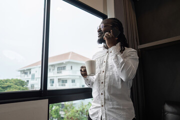African American man using have call by phone while drink coffee by the window in the living room