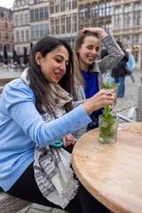 Two friends share a light-hearted moment at an outdoor cafe, one stirring a refreshing mojito. They're seated against the backdrop of a bustling city square, with historic buildings in the distance
