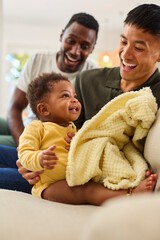 Same Sex Male Couple Or Friends Playing With Baby Sitting On Sofa At Home With Blanket Together