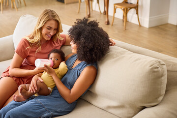 Same Sex Female Couple Or Friends Feeding Baby Girl With Bottle Sitting On Sofa At Home Together