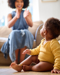 Smiling Baby Girl Learning To Crawl On Floor At Home With Mother Encouraging In Background