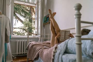 Woman reading a book in bed, lying on her stomach smiling happy and relaxed on a leisure day at home